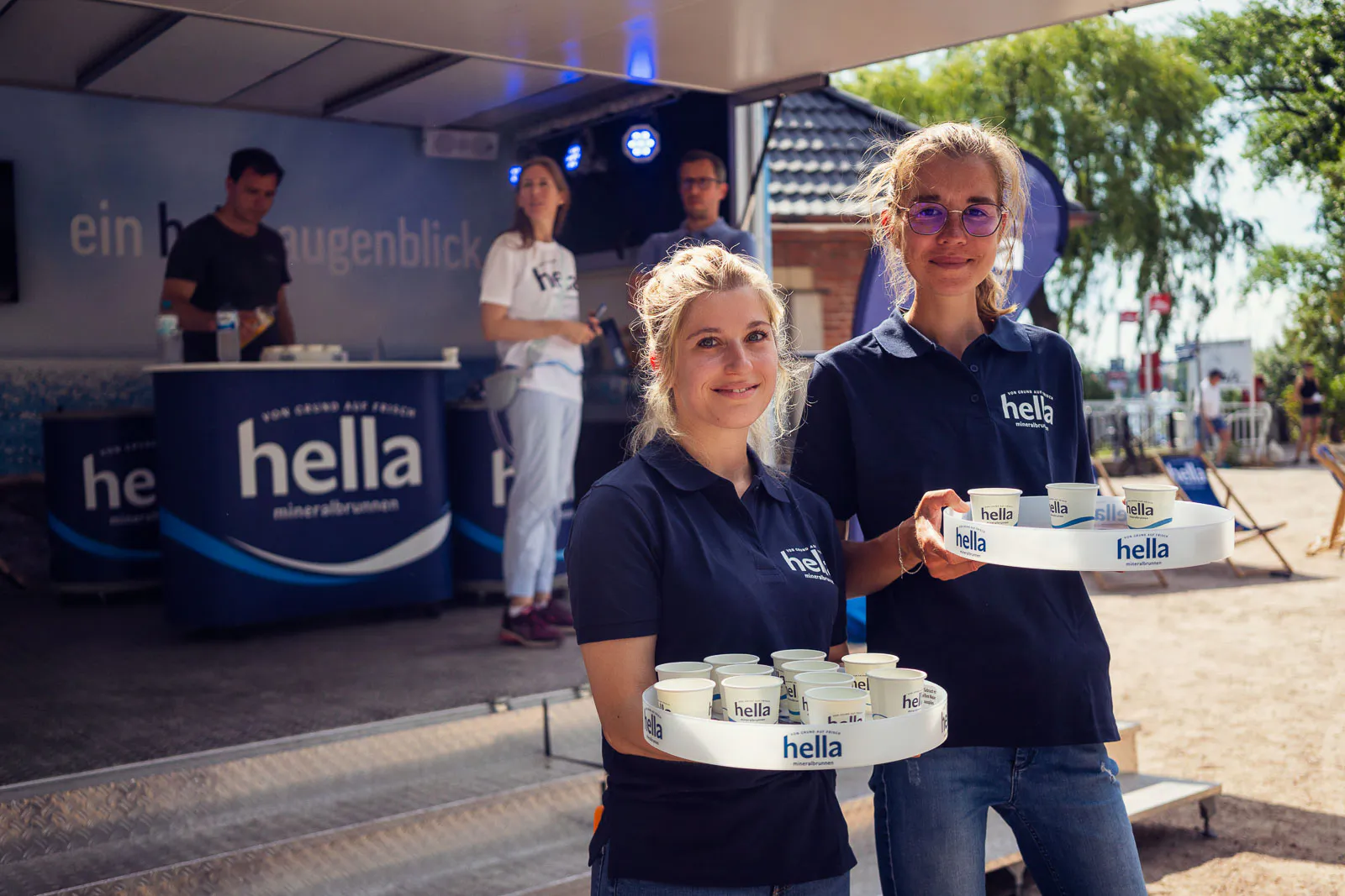 Zwei Frauen in Hella-Poloshirts halten Tabletts mit Getränkebechern vor einem sonnigen Getränkestand.