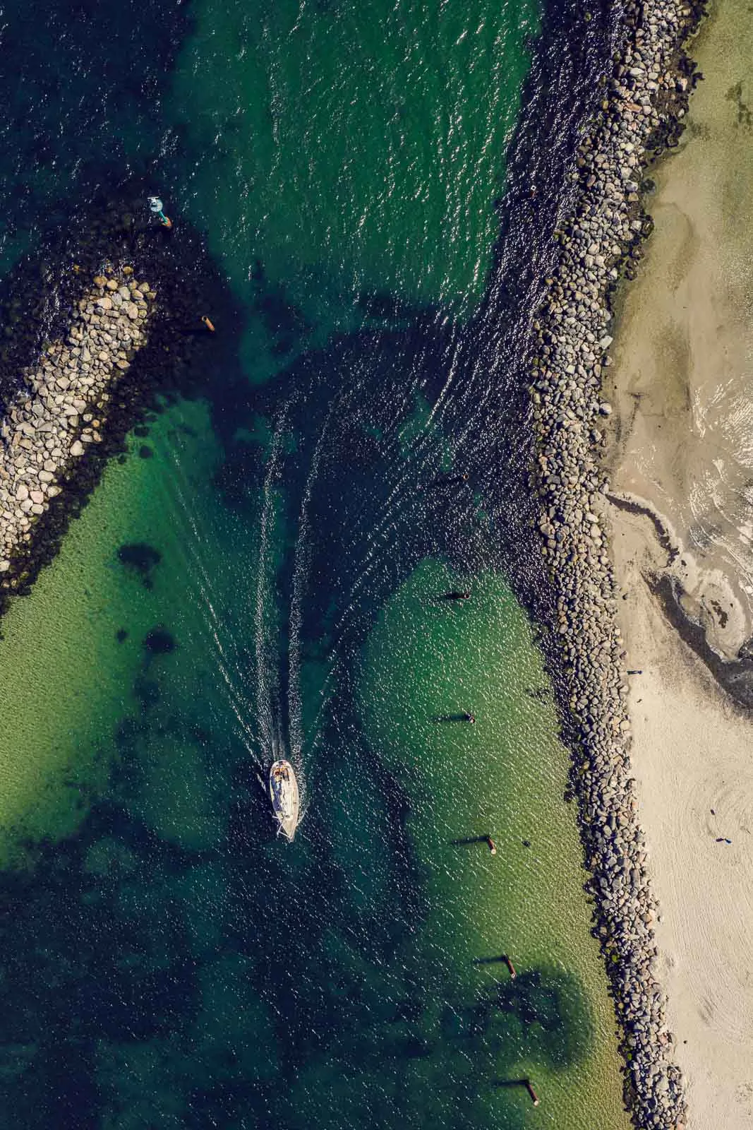 Luftaufnahme eines Boots mit Kielwasser im grünen Meer zwischen Steinmolen und Sandstrand.