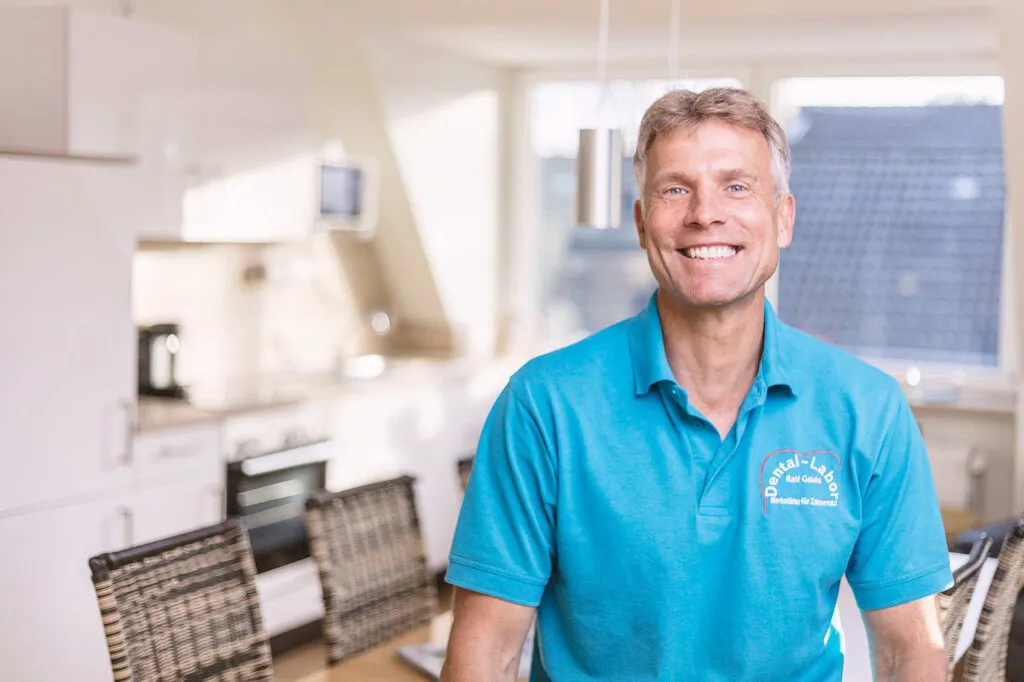 Lächelnder Mann im türkisfarbenen Poloshirt des Dental-Labors Ralf Gaida in einer hellen Küche mit Esstisch.