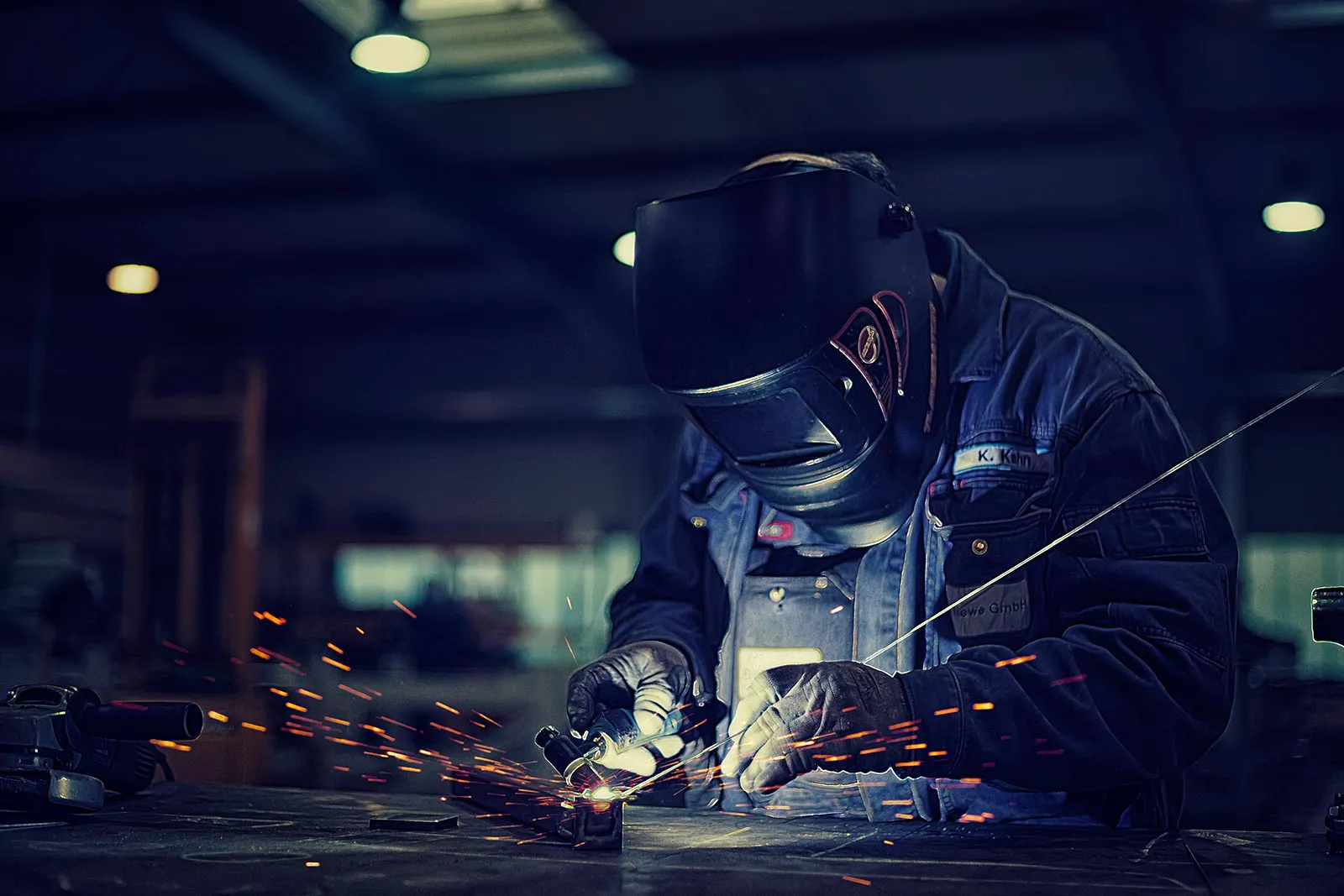 Schweißer mit Schutzhelm bearbeitet ein Metallstück am Werktisch; Funken leuchten in der dunklen Industriehalle.