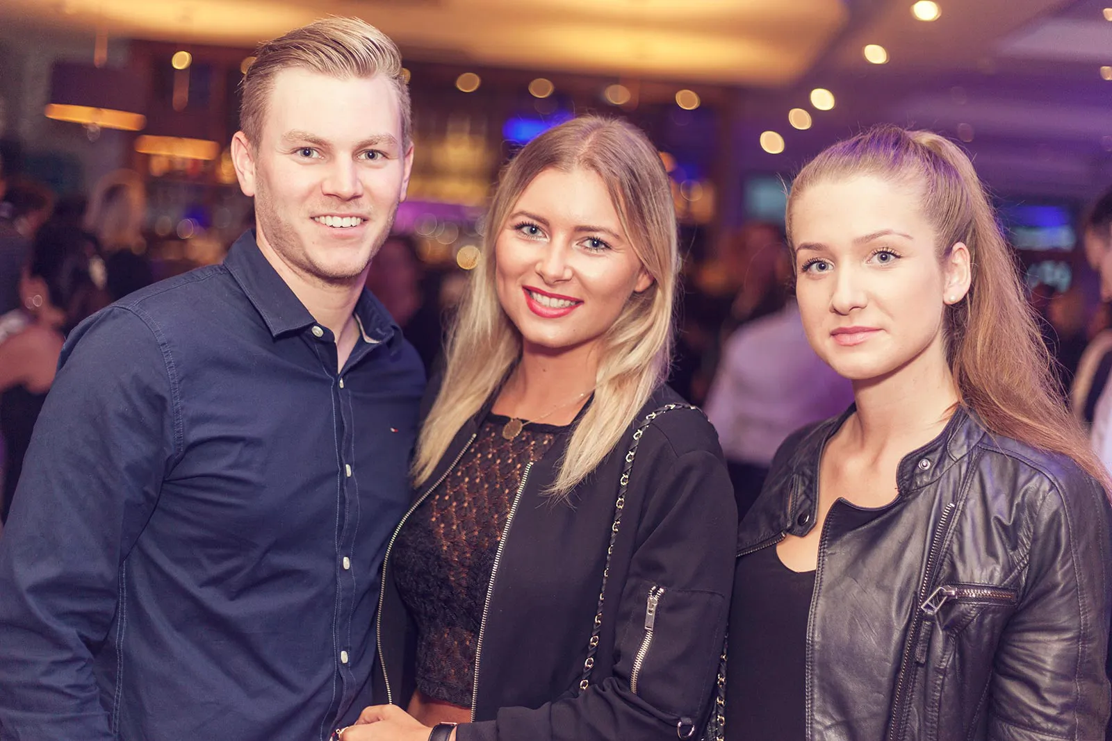 Ein Mann und zwei Frauen posieren bei warmem Licht in einer Bar, weitere Gäste stehen im Hintergrund.