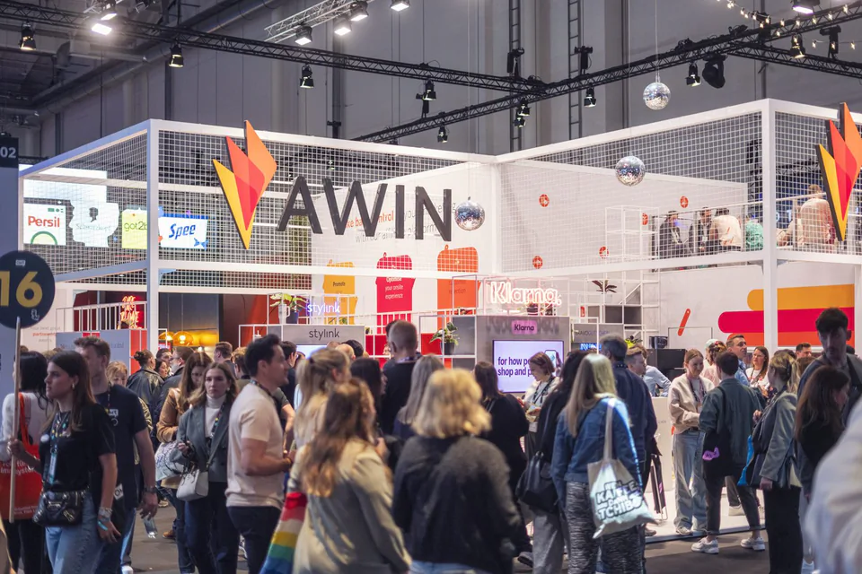 Zahlreiche Messebesucher vor dem zweistöckigen Awin-Stand mit großem Logo und Discokugeln in beleuchteter Messehalle.