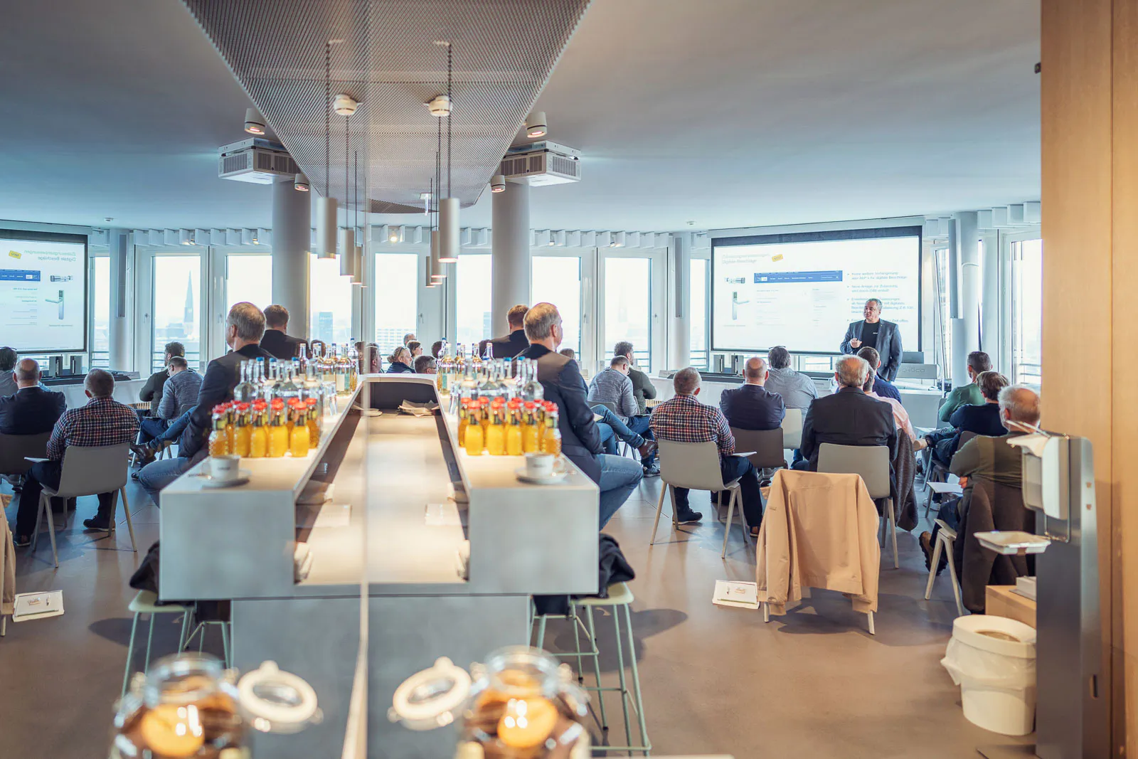 Vortrag beim Schüco-Event: Publikum vor einer Leinwand im hellen Raum, Getränkeflaschen an verspiegelter Theke.