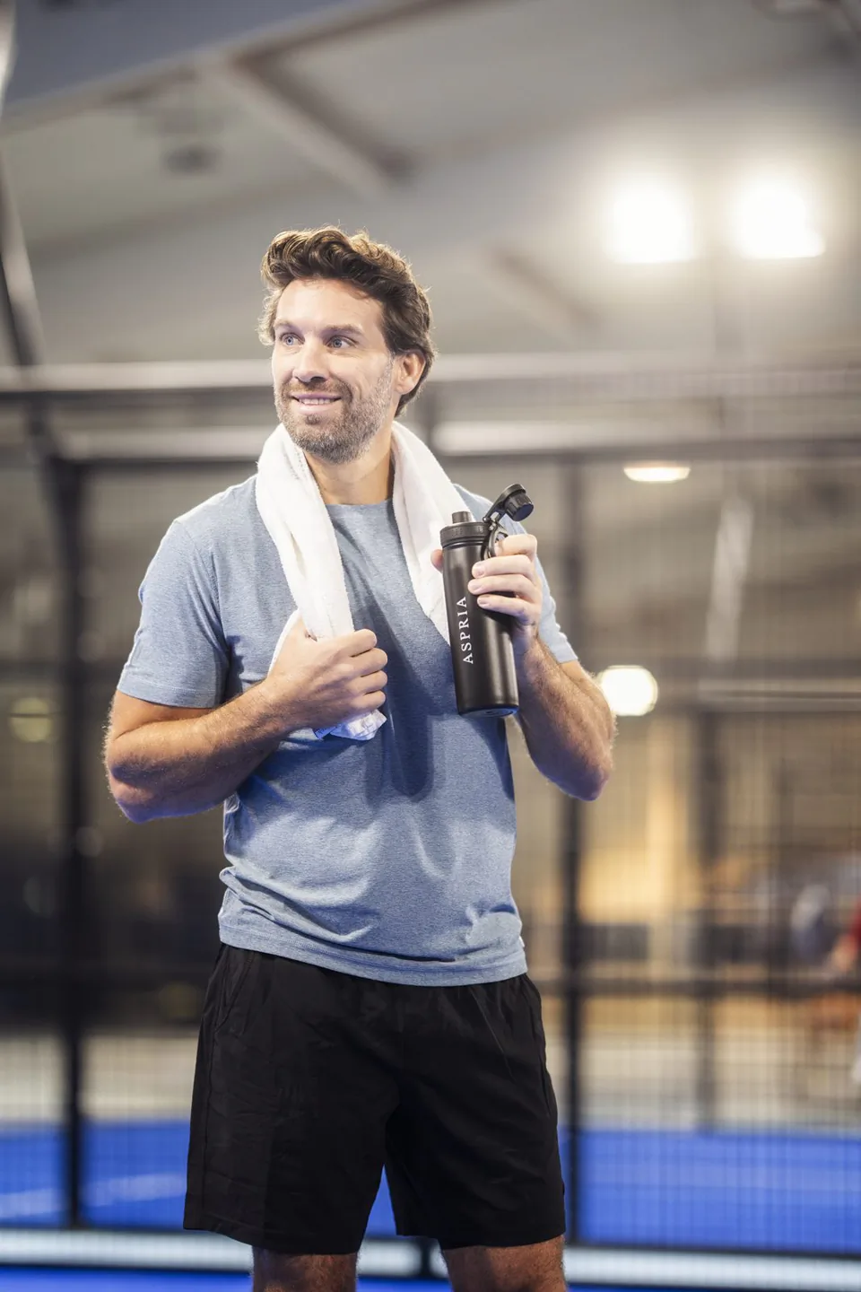 Mann mit Handtuch um den Hals und schwarzer Aspria-Trinkflasche auf einem blau beleuchteten Indoor-Padelplatz.