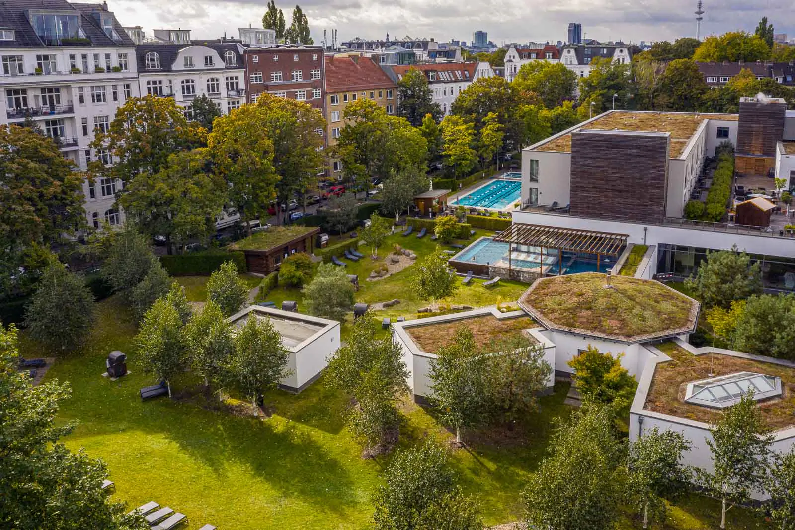 Drohnenansicht der Aspria-Anlage mit Außenpool, begrünten Dächern und Garten zwischen städtischen Wohnhäusern.
