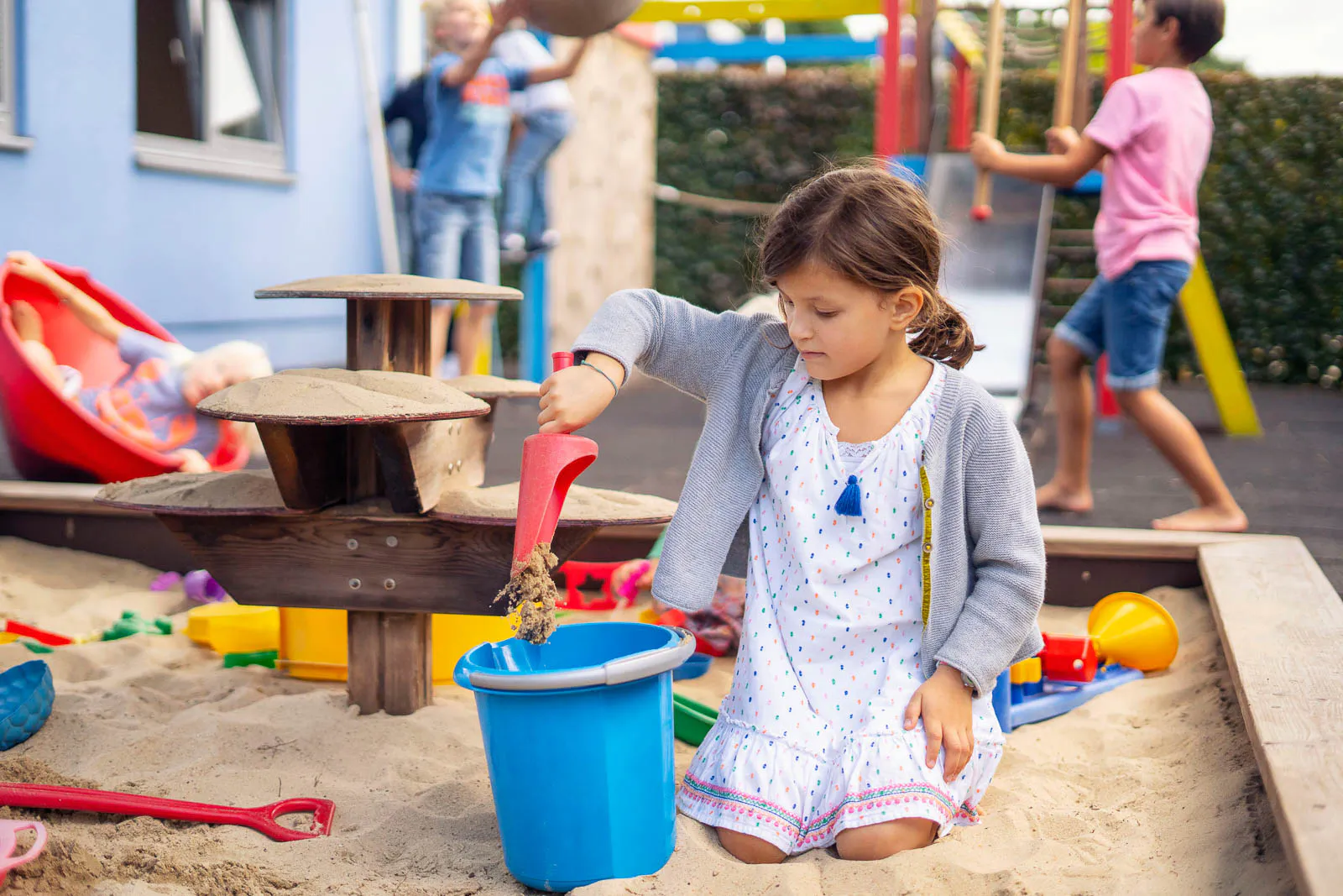 Ein Mädchen schaufelt Sand in einen blauen Eimer im Sandkasten, weitere Kinder spielen im Hintergrund.