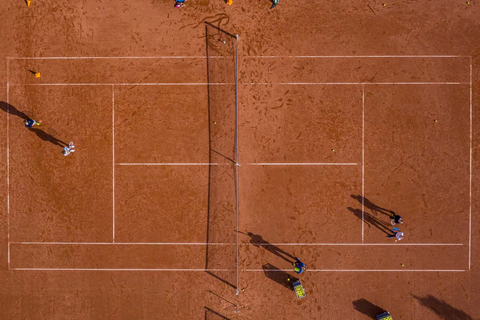 Drohnenansicht eines roten Tennisplatzes mit Spielern an den Grundlinien und langen Schatten im Sonnenlicht.