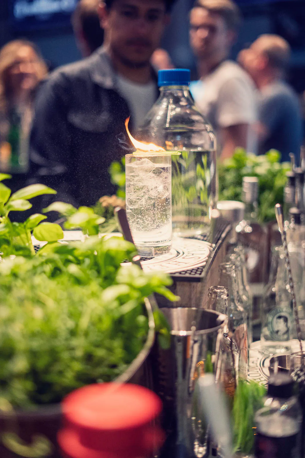 Brennender Cocktail im hohen Glas auf einer Bar, umgeben von frischen Kräutern und Flaschen, dahinter Eventgäste.
