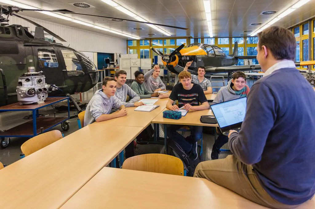 Schüler sitzen vor einem Lehrer mit Laptop in einer hellen Werkhalle mit Hubschrauber und Propellerflugzeug.