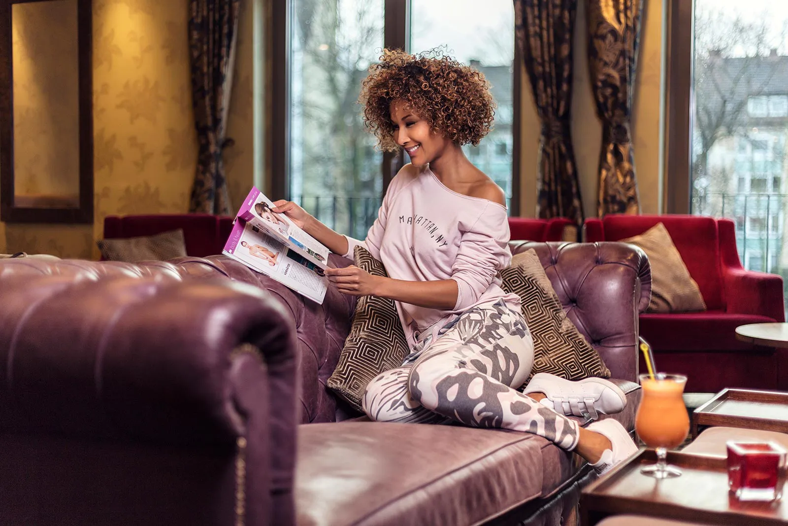 Marie Amière liest lächelnd auf einem Ledersofa in einer Lounge; auf dem Tisch steht ein orangefarbener Drink.