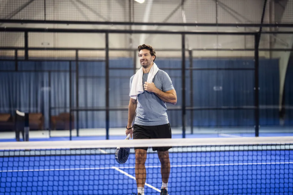 Mann mit Padelschläger und Handtuch um den Hals steht am Netz auf einem blauen Padelplatz in einer beleuchteten Halle.