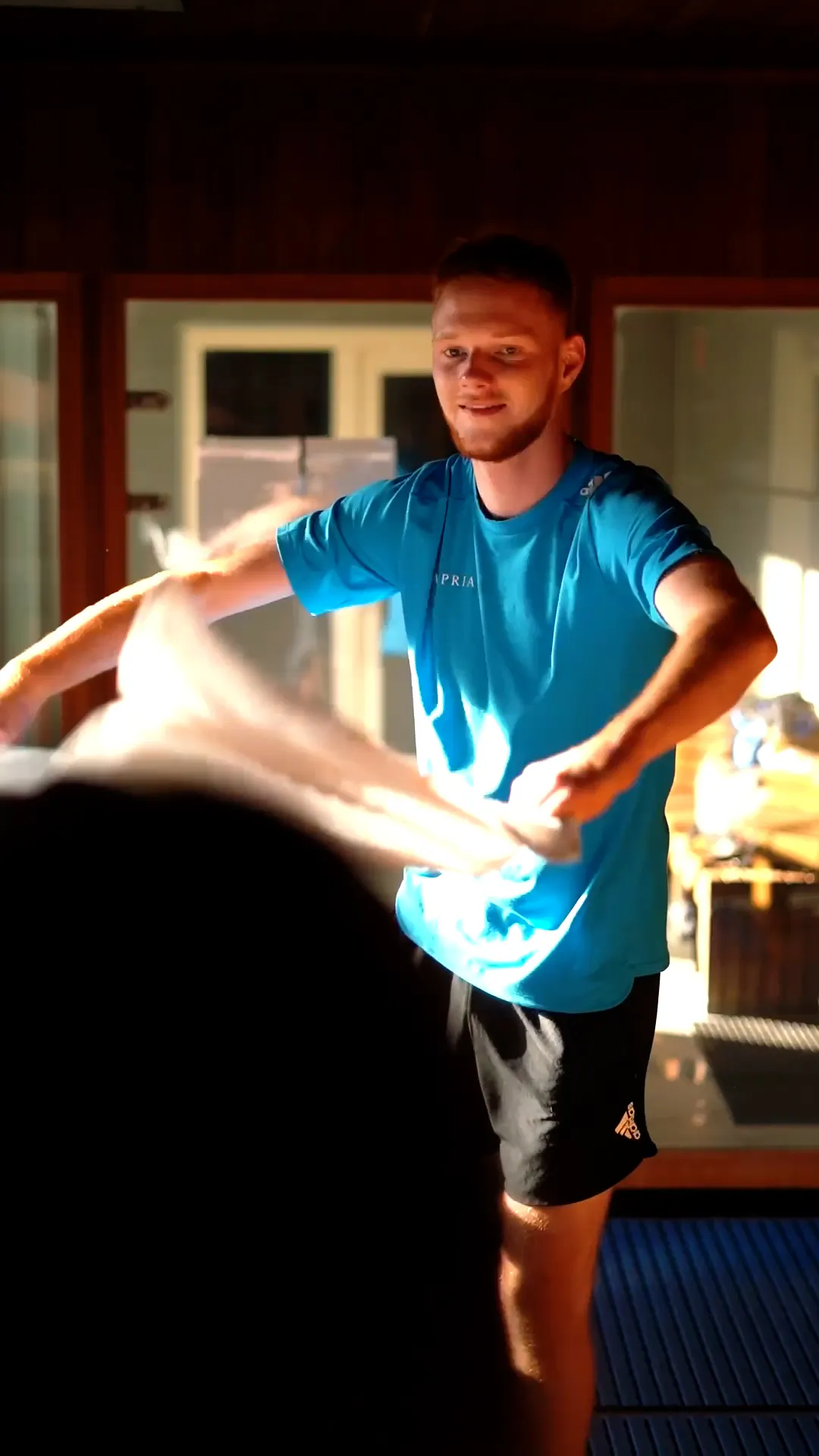 Mann im blauen Shirt wedelt mit einem Handtuch in einer holzverkleideten Sauna bei warmem Licht.