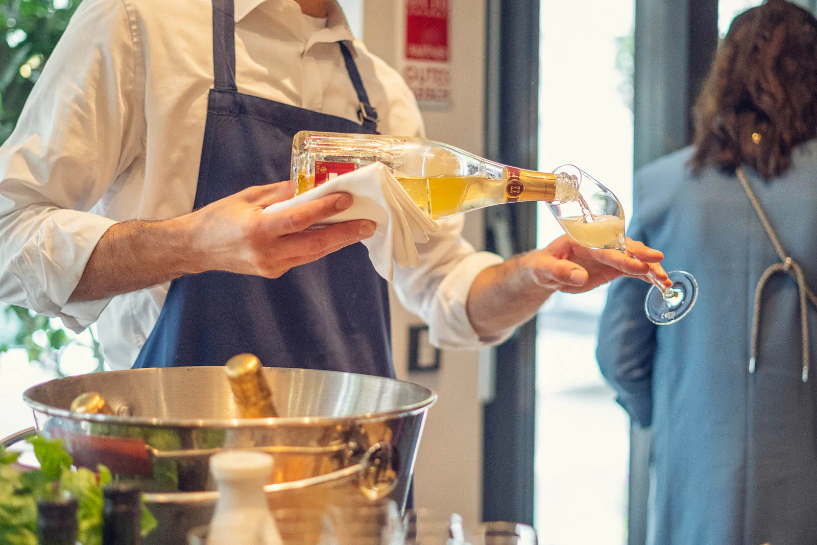 Servicekraft mit dunkler Schürze schenkt im hellen Restaurant Weißwein ins Glas, davor steht ein Weinkühler.