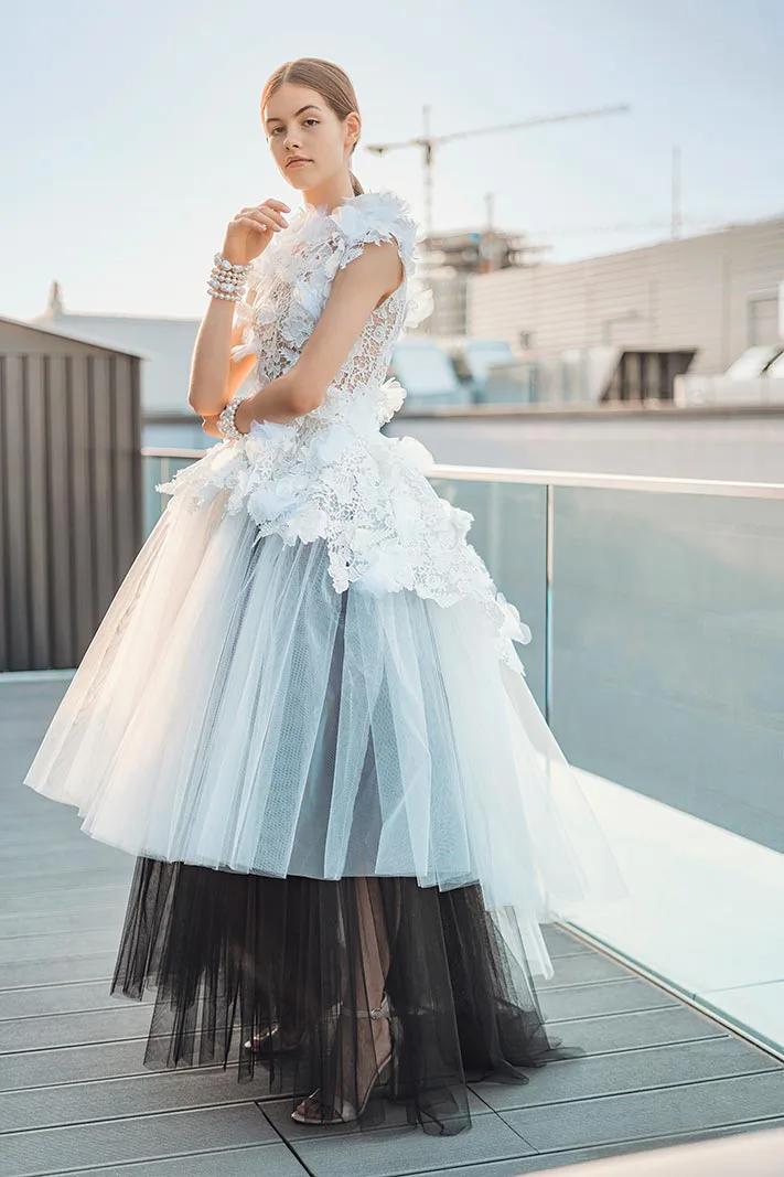 Model in hellblauem Tüllkleid mit Blütenapplikationen und schwarzem Unterrock posiert im Sonnenlicht auf einer Terrasse.