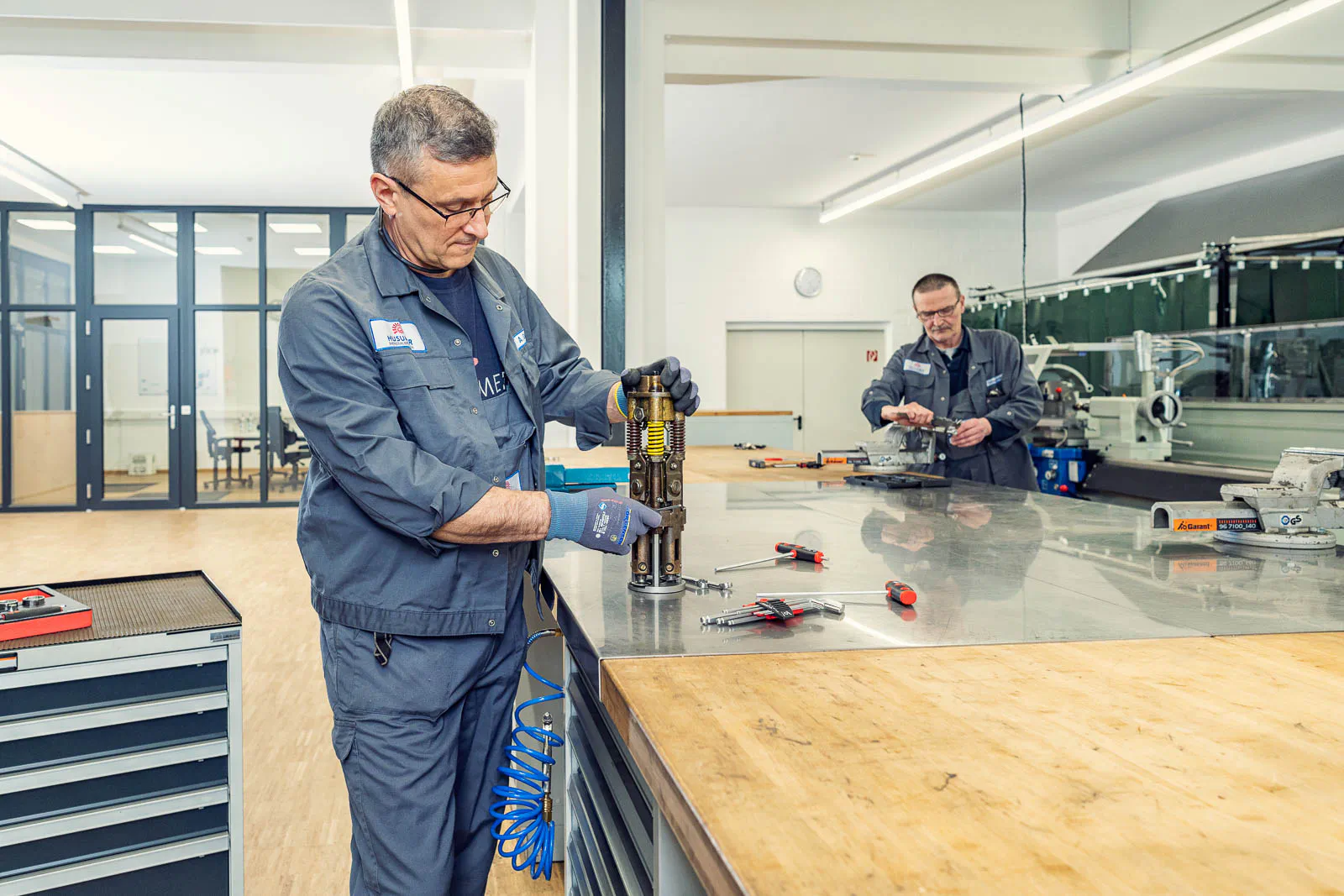 Zwei Mitarbeiter in grauer Arbeitskleidung bearbeiten Metallteile an Werkbänken in einer hell beleuchteten Werkstatt.