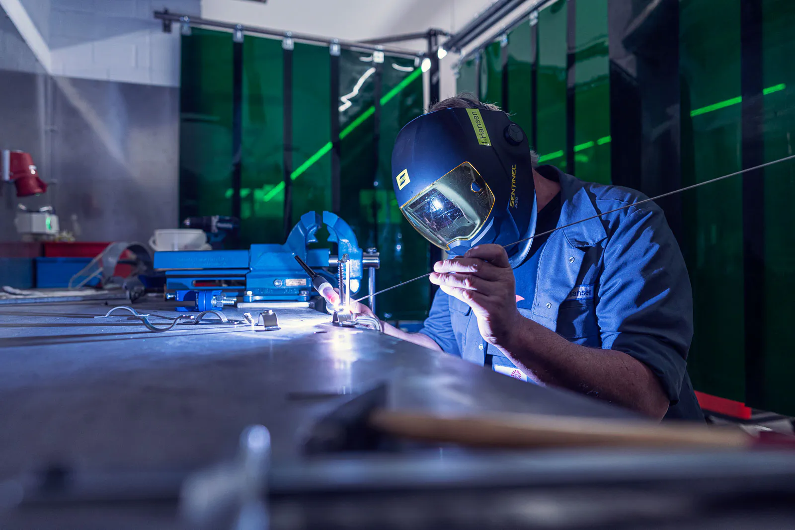 Mitarbeiter mit Schweißhelm schweißt ein Metallteil an einer Werkbank, beleuchtet vom hellen Lichtbogen.
