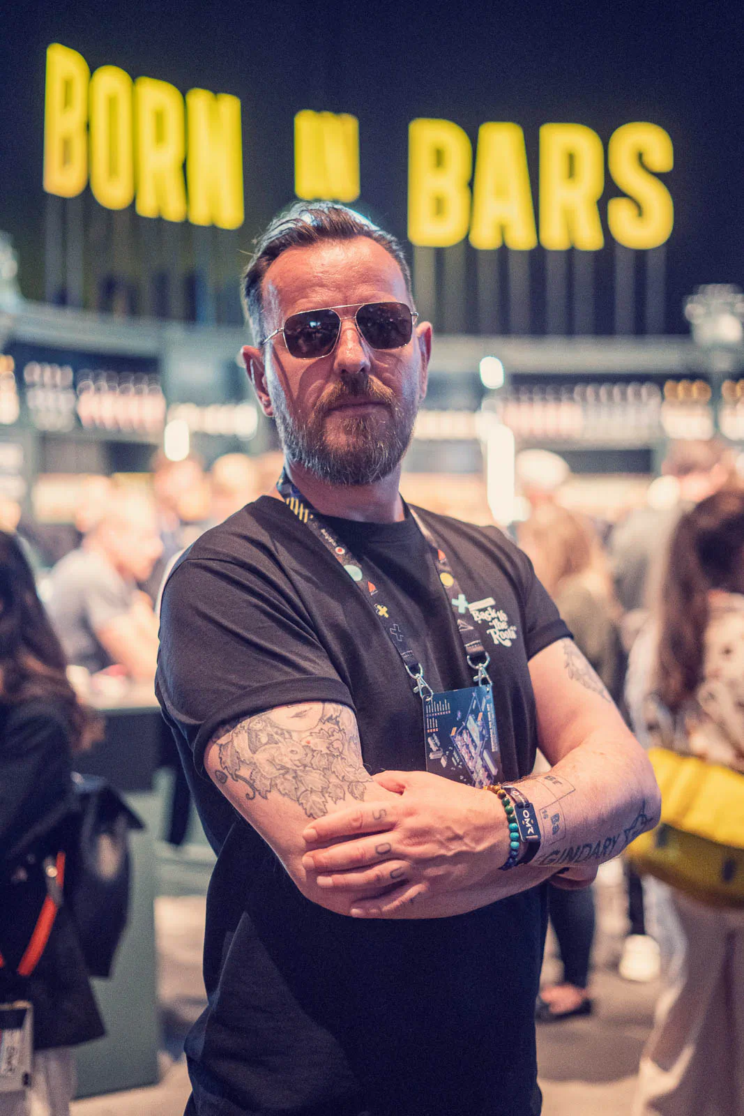 Tätowierter Mann mit Sonnenbrille und verschränkten Armen vor einer Bar mit leuchtendem Schriftzug BORN IN BARS.