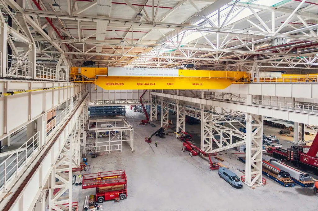 Gelber Konecranes-Brückenkran in heller Industriehalle mit weißen Stahlträgern, Hebebühnen und Fahrzeugen.