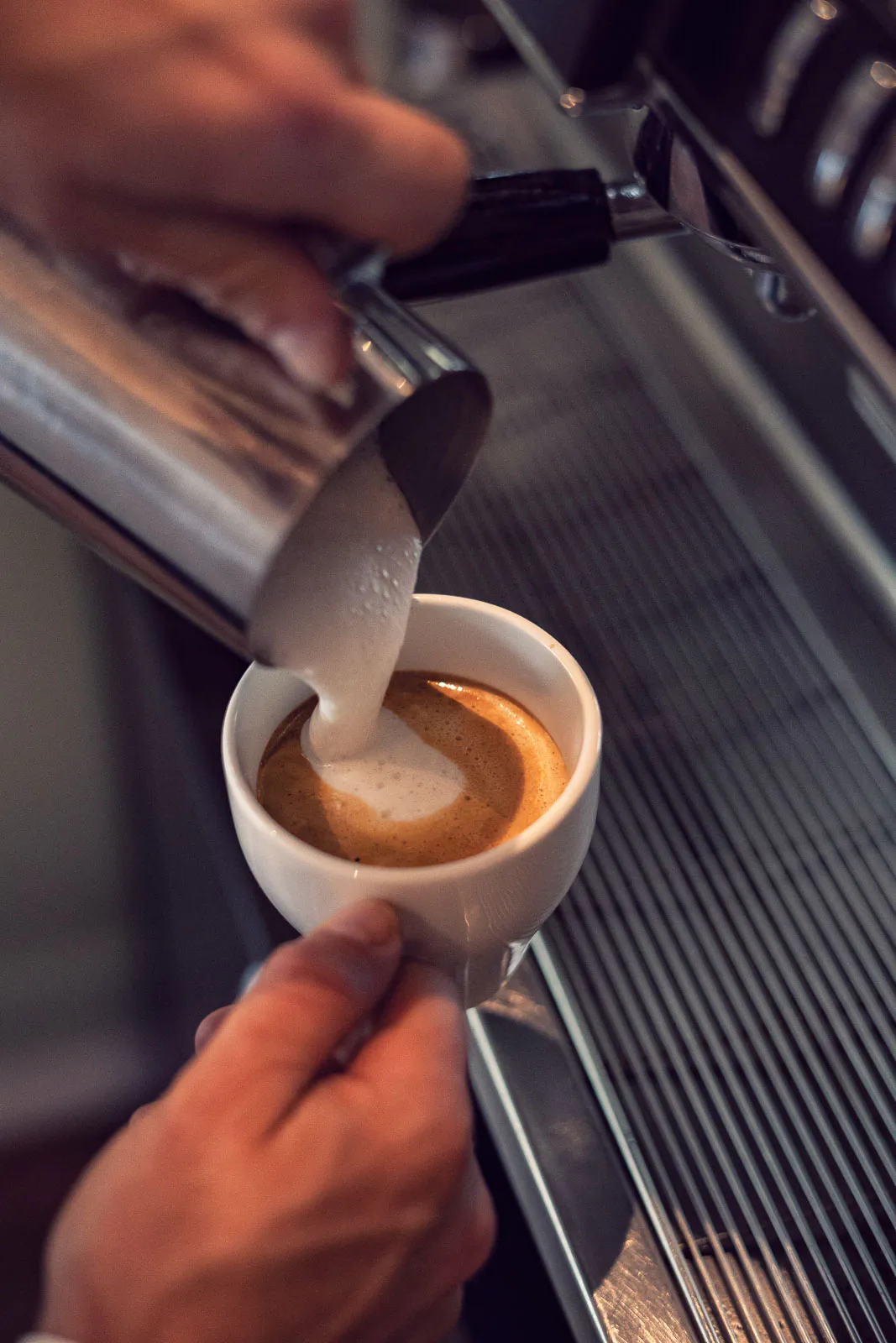 Hände gießen Milchschaum aus einem Edelstahlkännchen in eine weiße Kaffeetasse an einer Espressomaschine.
