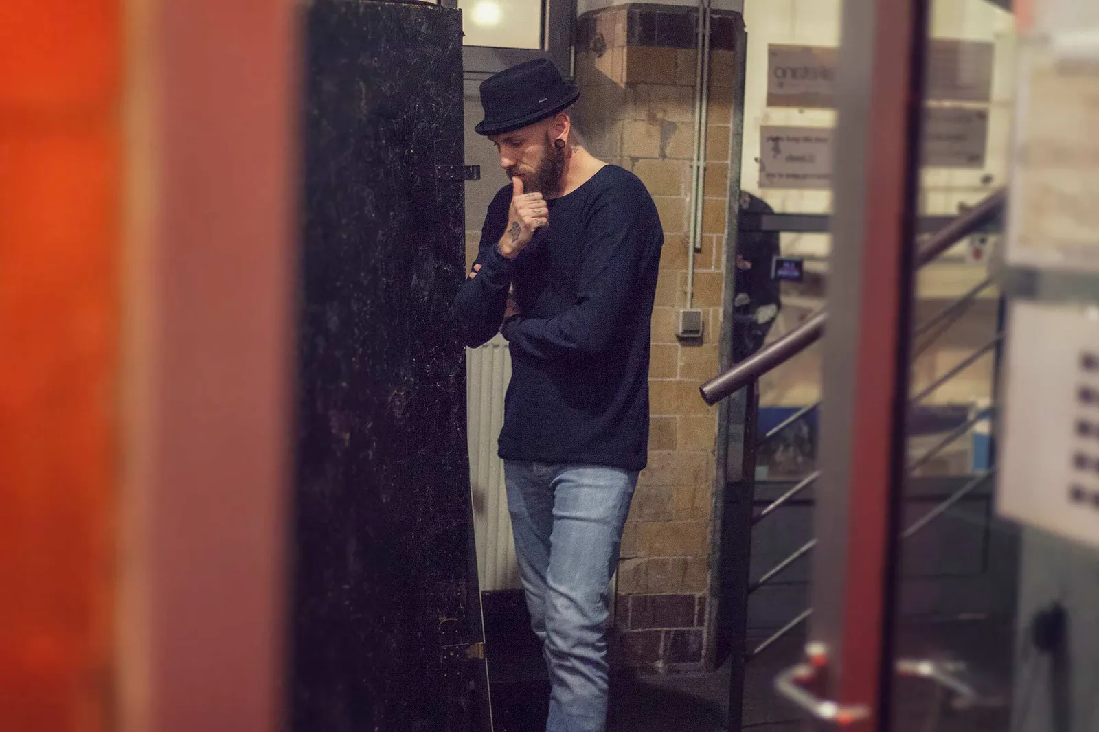 Dennis Grundt steht mit schwarzem Hut und Hand am Kinn neben einer Tür im Treppenhaus.
