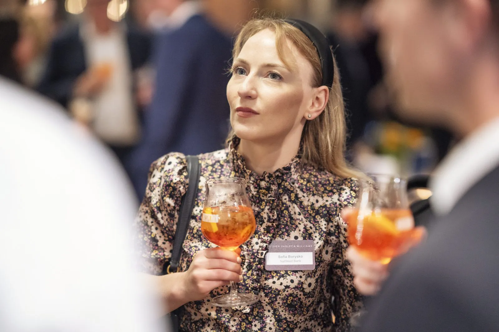 Frau im geblümten Kleid mit Namensschild und orangefarbenem Drink zwischen Gästen bei warmem Licht im Innenraum.