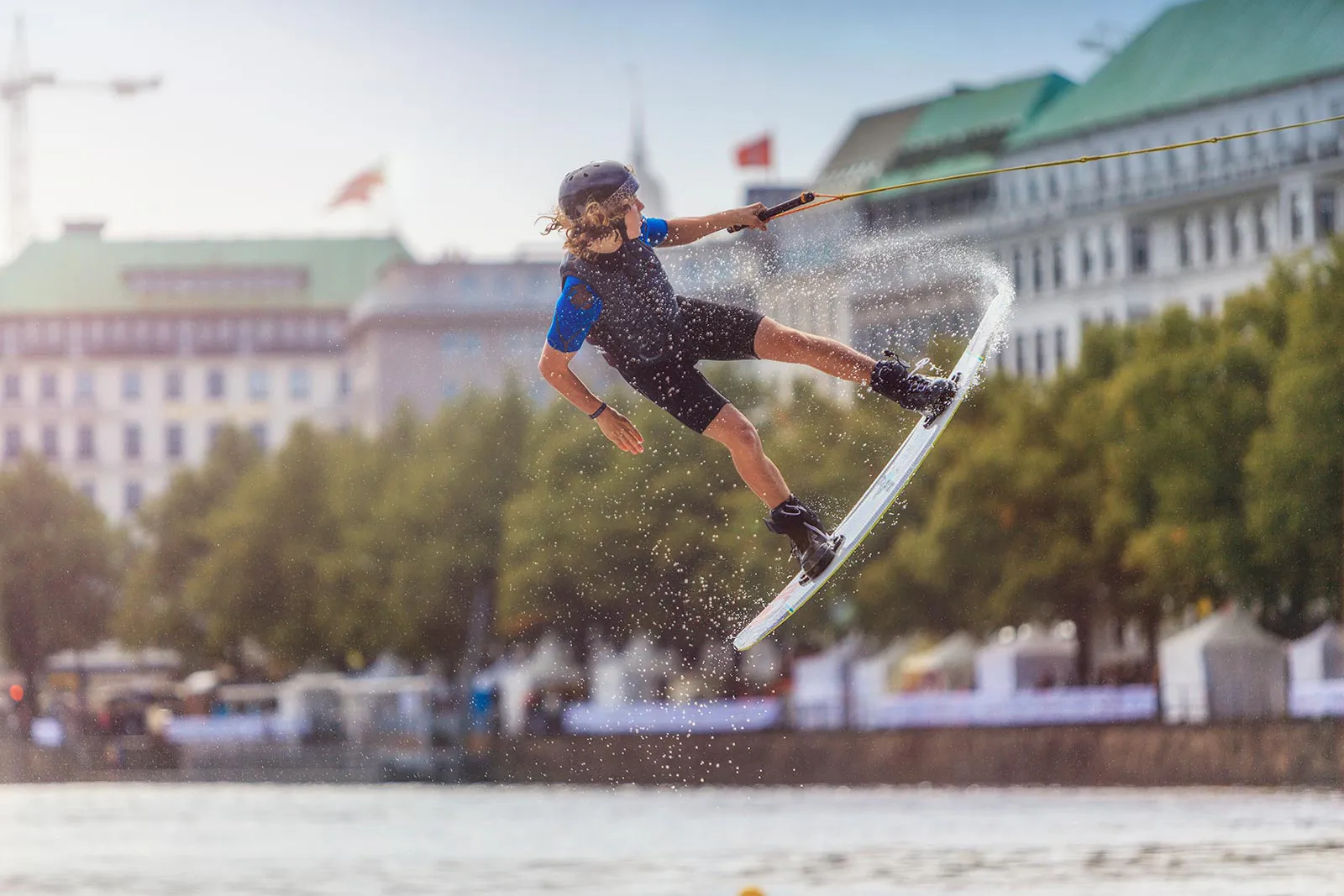 Wakeboarder mit Helm springt am Zugseil über das Wasser, vor einer sonnenbeschienenen Uferpromenade mit Gebäuden.