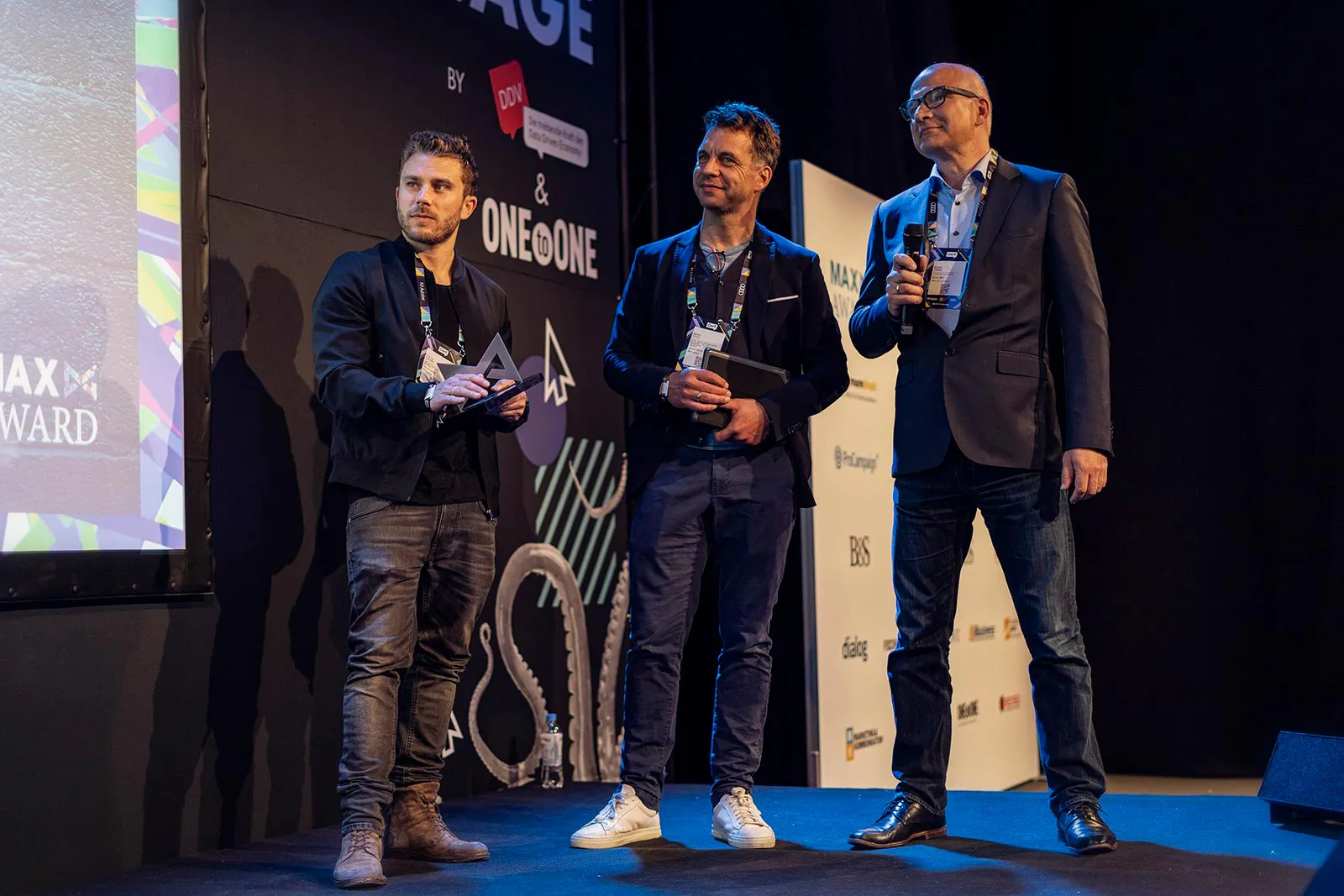 Drei Männer auf blau beleuchteter Bühne beim MAX Award, einer spricht ins Mikrofon, zwei halten Unterlagen.