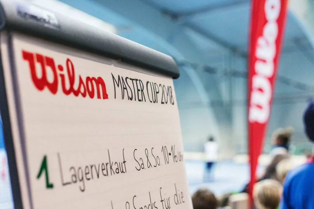 Aufsteller zum Wilson Master Cup mit Hinweis auf Lagerverkauf, dahinter Menschen und rote Wilson-Fahne in einer Sporthalle.