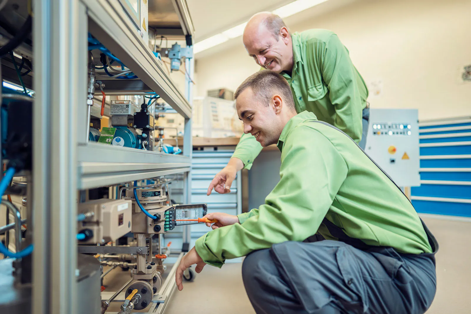 Zwei Männer in grüner Arbeitskleidung prüfen eine technische Anlage in einer hellen Werkstatt der Raffinerie Heide.