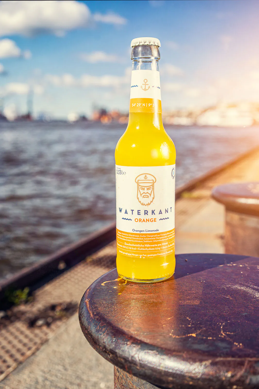 Eine Flasche Waterkant Orangenlimonade steht im warmen Sonnenlicht auf einem Poller am Hafenkai.