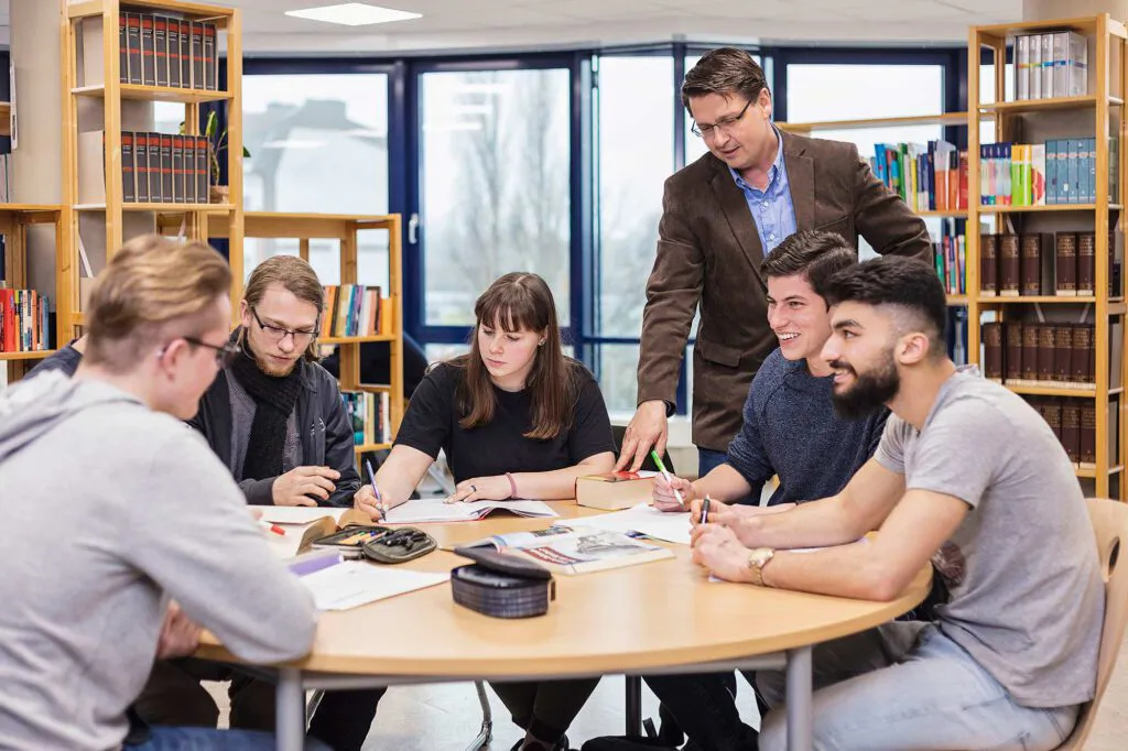Sechs Personen lernen und besprechen Unterlagen an einem runden Tisch in einer hellen Bibliothek.