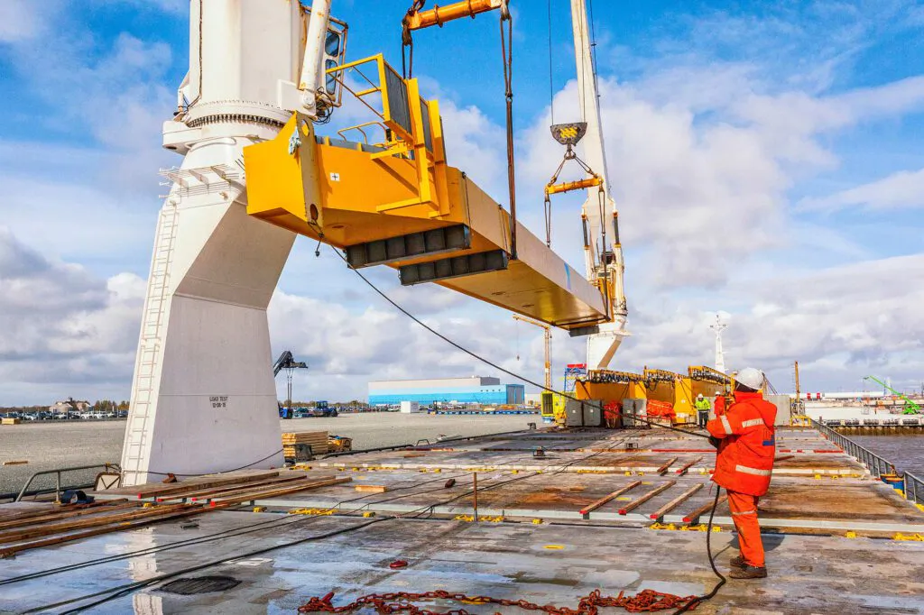Arbeiter in orangefarbener Schutzkleidung auf nassem Schiffsdeck unter einem Kran mit gelber Last im Sonnenlicht.