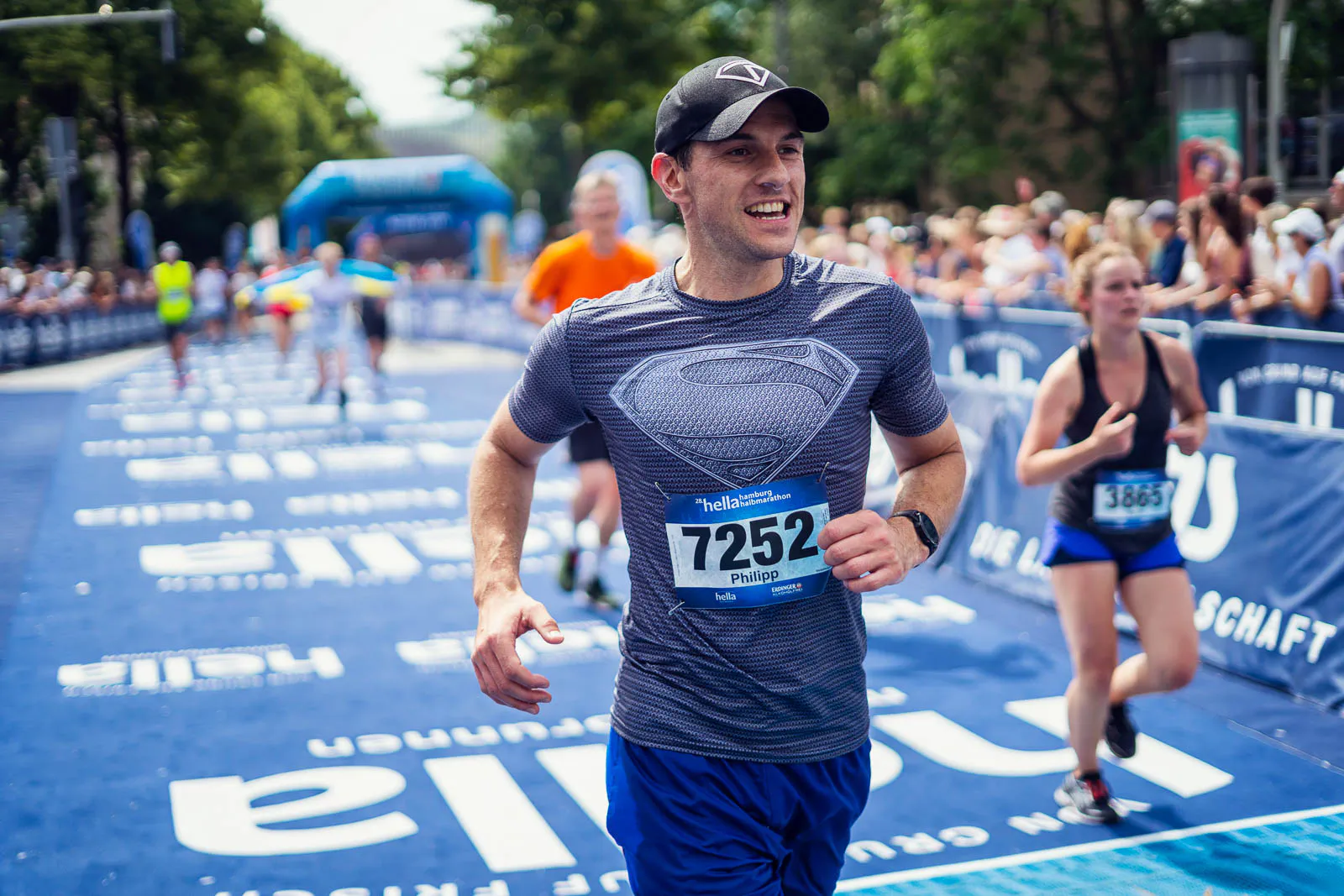 Läufer mit Superman-Shirt und Startnummer 7252 auf blauer Laufstrecke, dahinter Läuferin und Zuschauer im Sonnenlicht.