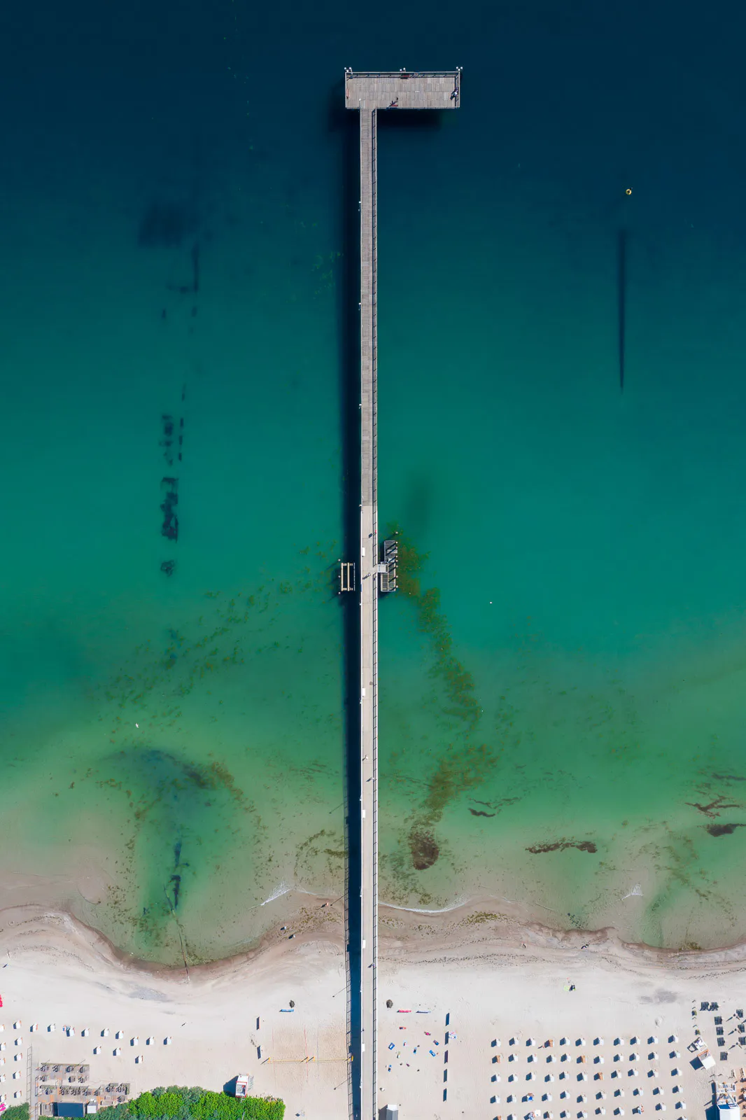 Luftaufnahme einer langen Seebrücke im türkisblauen Meer mit Sandstrand und Strandkörben bei Sonnenlicht.