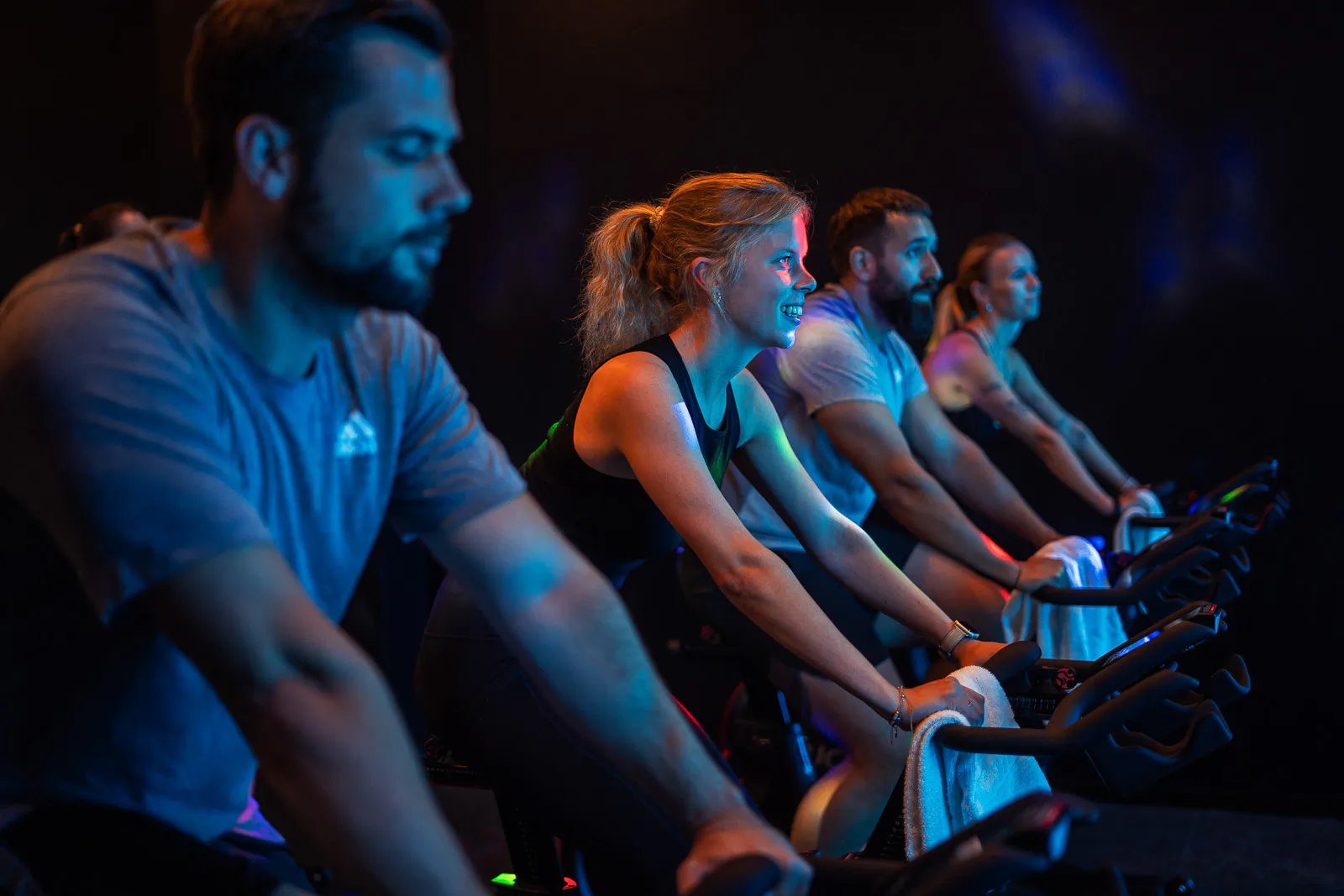 Vier Personen trainieren auf Indoor-Cycling-Rädern in einem dunklen Raum mit blauem und orangefarbenem Licht.