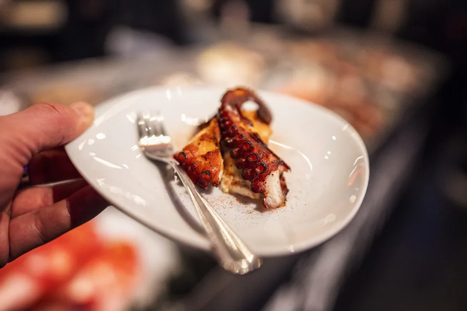 Eine Hand hält einen weißen Teller mit gebratenem Oktopus und einer Gabel vor einem unscharfen Buffet.