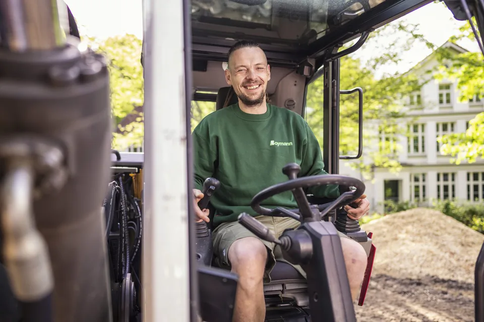 Lächelnder Mann im grünen Boymann-Pullover sitzt am Steuer einer Baumaschine, dahinter Bäume und ein Gebäude im Tageslicht.