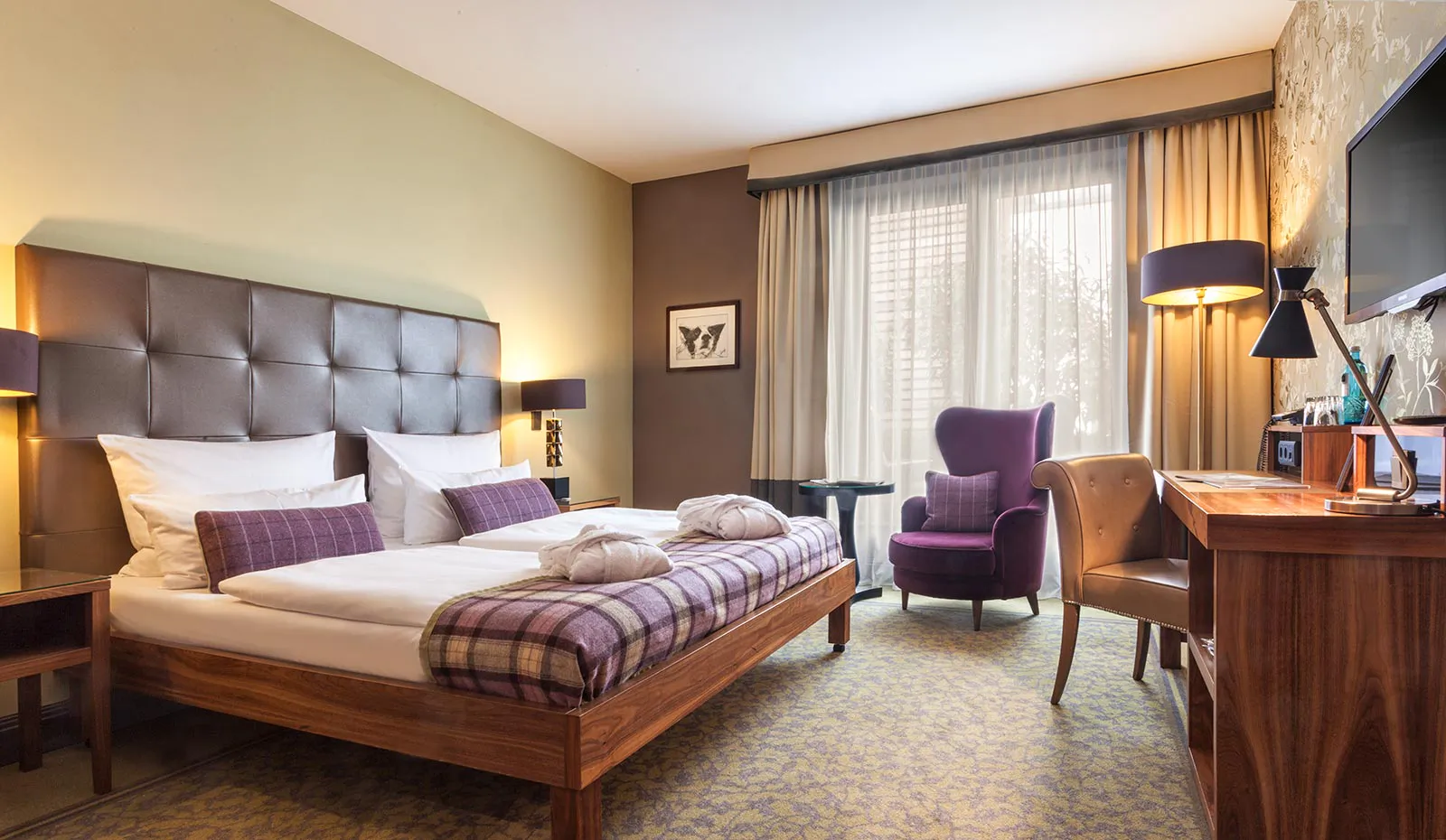 Hotelzimmer mit Doppelbett, karierten Decken, violettem Sessel und Holzschreibtisch im warmen Tages- und Lampenlicht.