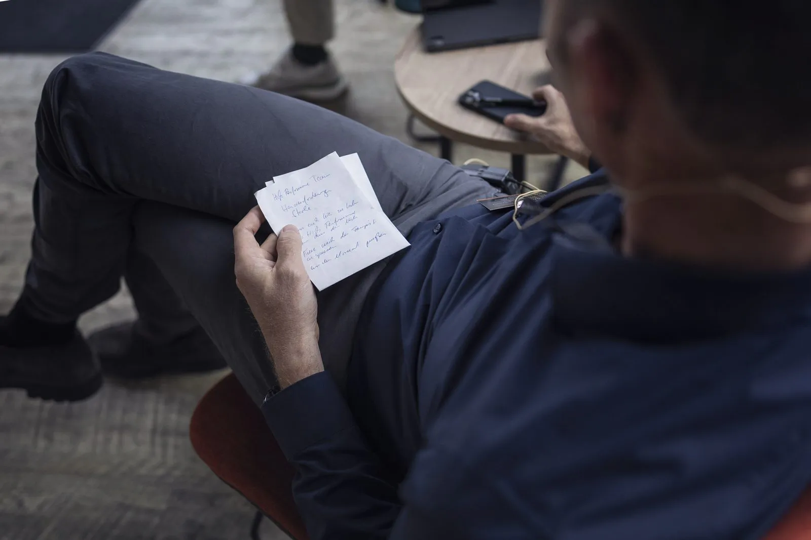 Sitzender Mann mit Headset-Mikrofon hält handschriftliche Notizen neben einem kleinen Tisch mit Smartphone.