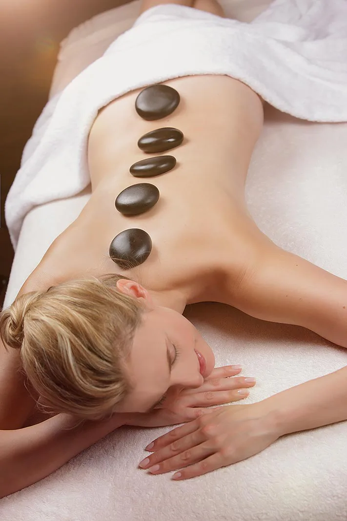 Frau liegt bei warmem Licht auf einer Spa-Liege, fünf dunkle Massagesteine auf dem Rücken, ein weißes Handtuch um die Hüfte.