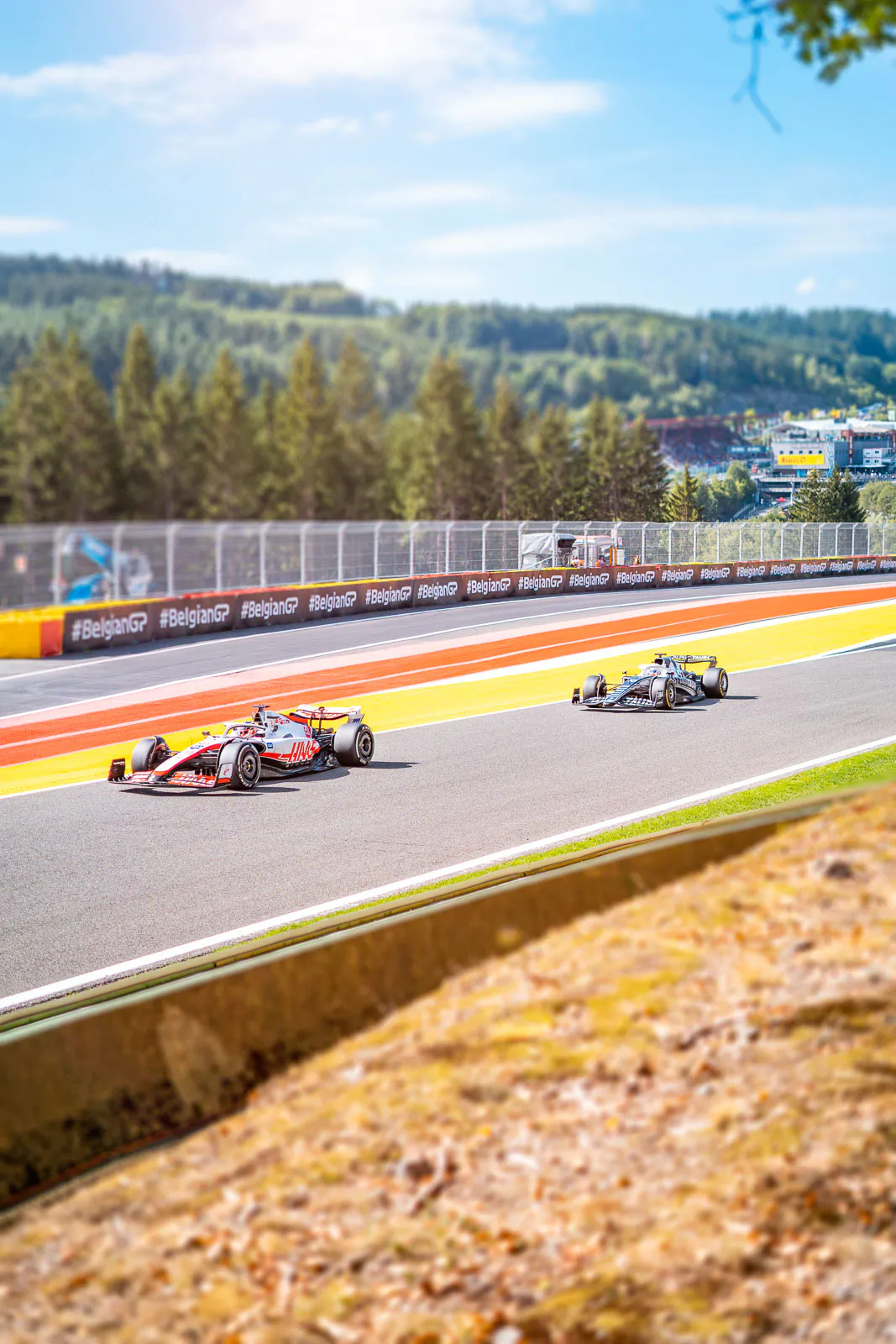 Zwei Formel-1-Rennwagen auf sonniger Rennstrecke mit rot-gelben Randstreifen und bewaldeten Hügeln im Hintergrund.