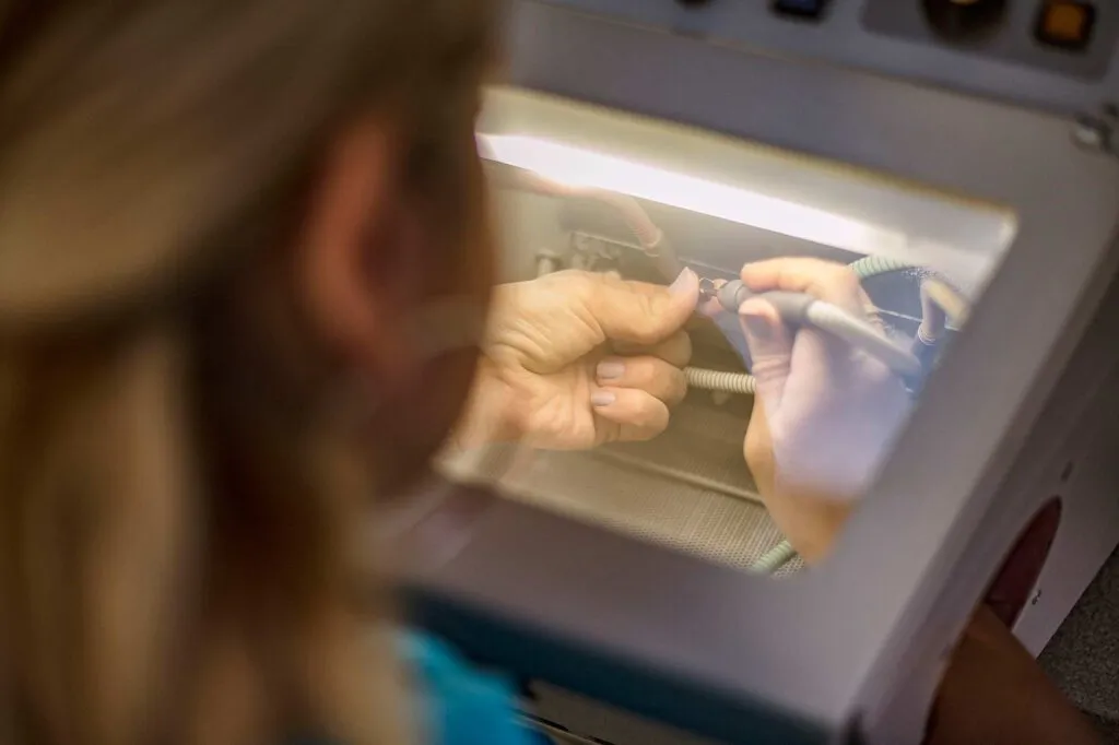 Person bearbeitet ein kleines Dentalteil mit einem Handgerät hinter der beleuchteten Schutzscheibe im Dentallabor.