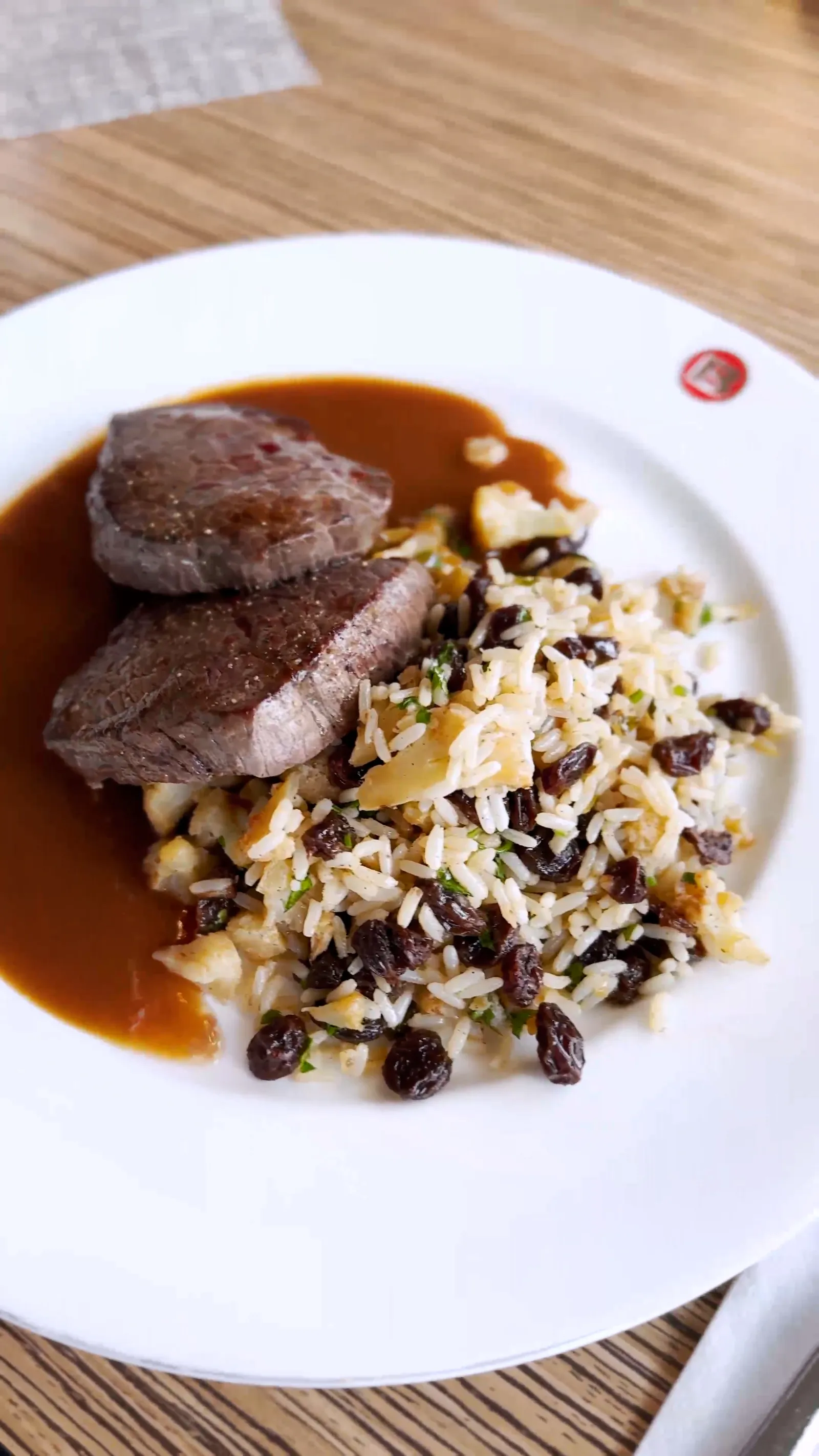 Zwei gebratene Fleischstücke mit dunkler Sauce und einer Reisbeilage auf einem weißen Teller auf einem Holztisch.