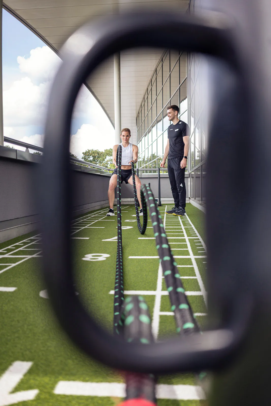 Frau trainiert mit Battle Ropes neben einem Mann auf einer sonnigen Außenterrasse mit Kunstrasen.