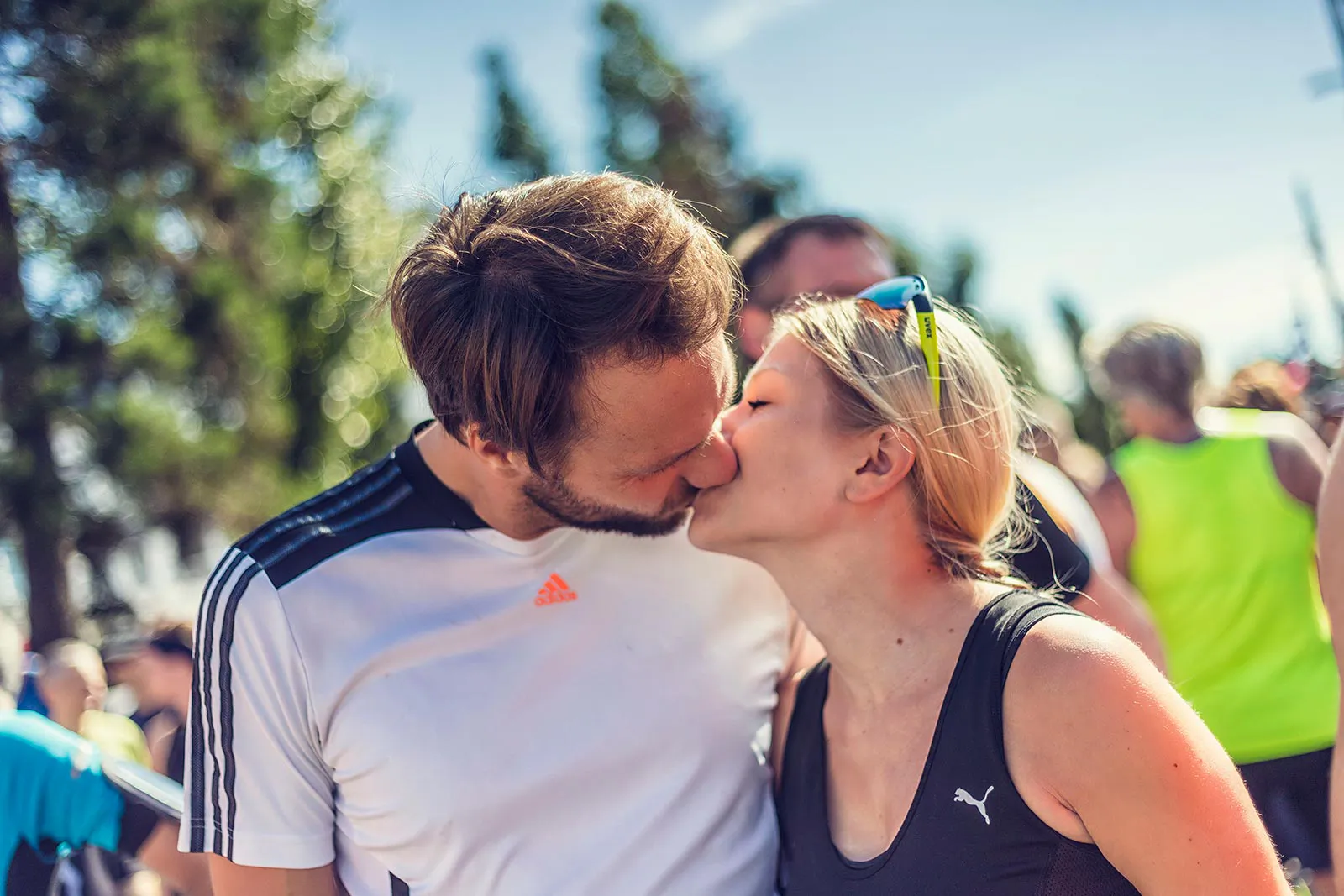 Ein Mann im weißen Sportshirt und eine Frau im schwarzen Sporttop küssen sich im Sonnenlicht zwischen Laufteilnehmern.