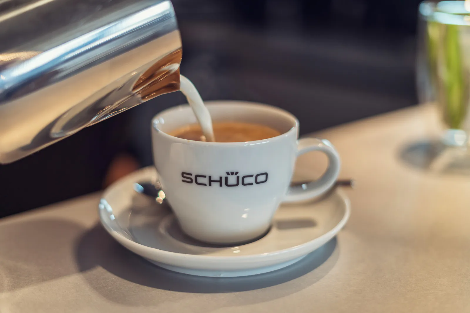 Eine helle Flüssigkeit wird aus einer Metallkanne in eine weiße Schüco-Tasse auf einer Untertasse mit Löffel gegossen.