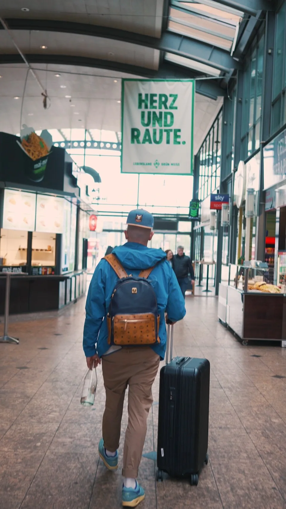Person mit blauer Jacke und Rucksack zieht einen Rollkoffer durch eine helle Halle mit Verkaufsständen.