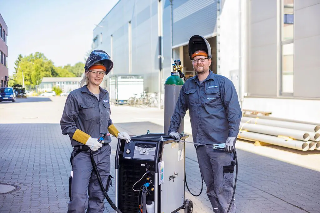 Zwei Personen mit hochgeklappten Schweißhelmen stehen bei Sonnenlicht mit einem Schweißgerät vor einer Industriehalle.