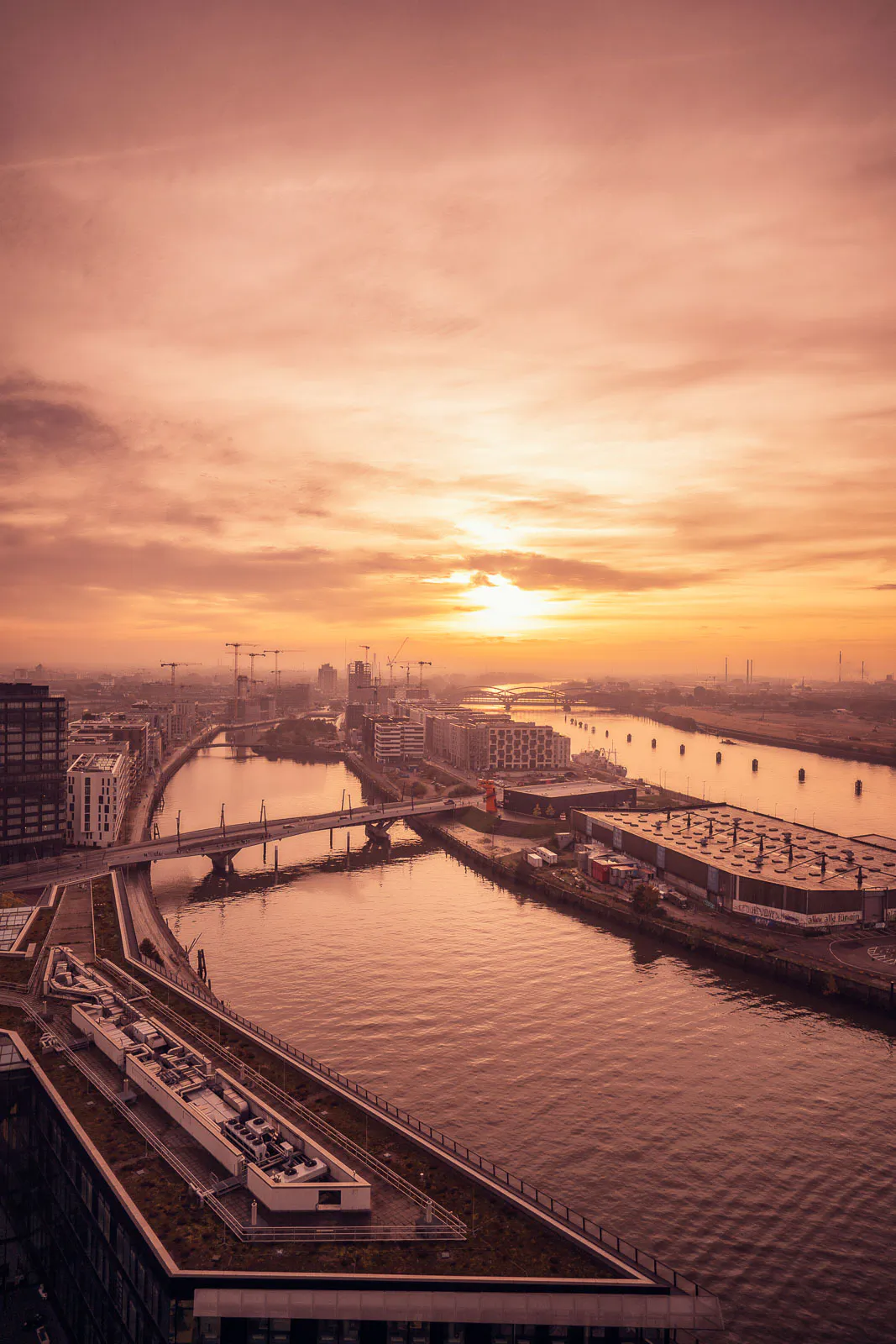 Hafenbecken mit Brücke, Kränen und Gebäuden im warmen Licht der tief stehenden Sonne unter orangefarbenem Himmel.