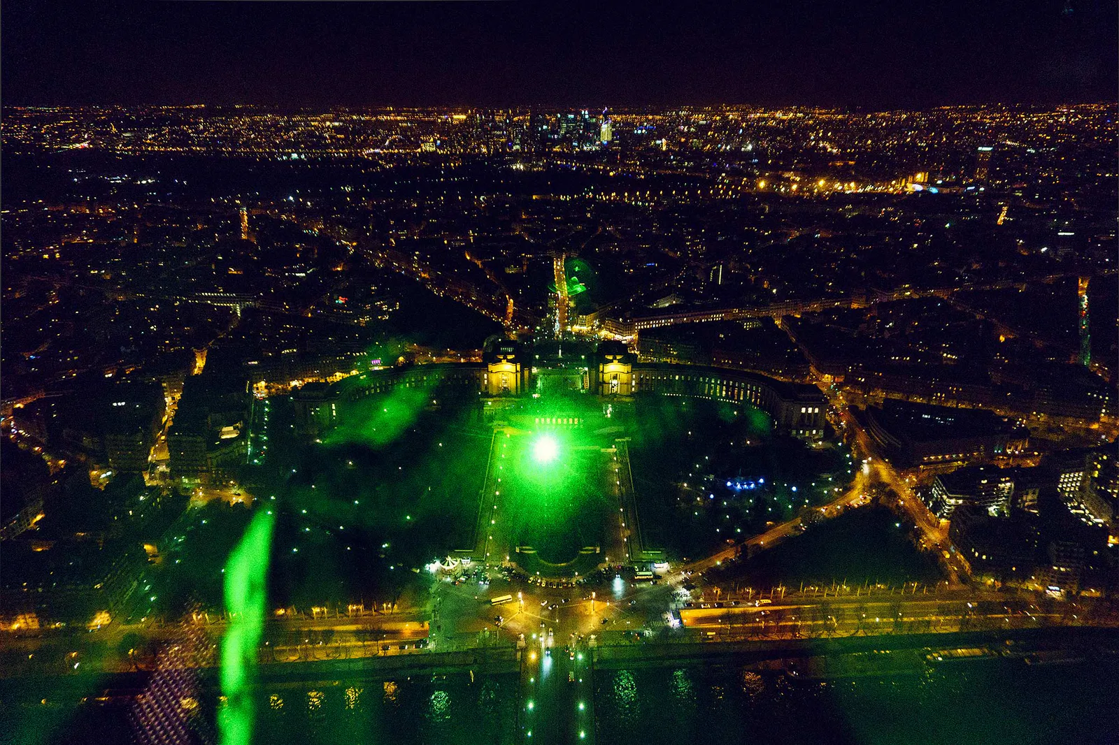 Nächtlicher Blick über Paris mit beleuchteten Straßen, Seine und grünen Lichtreflexen im Vordergrund.